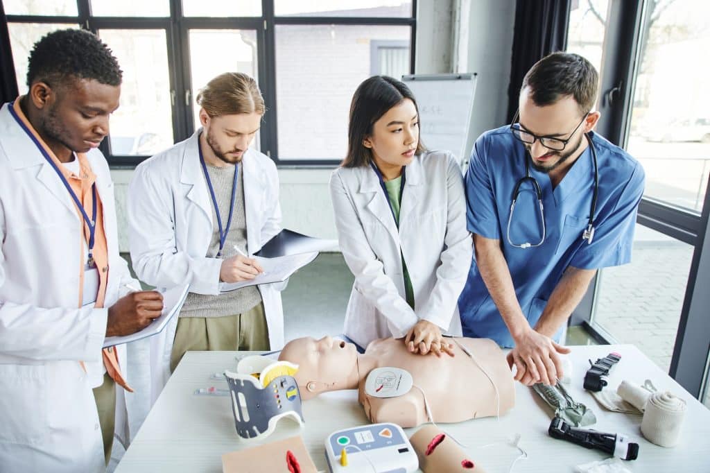 multiethnic students in white coats writing in notebooks near medical instructor assisting asian