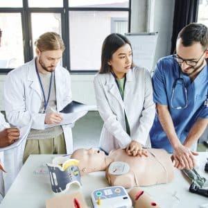multiethnic students in white coats writing in notebooks near medical instructor assisting asian
